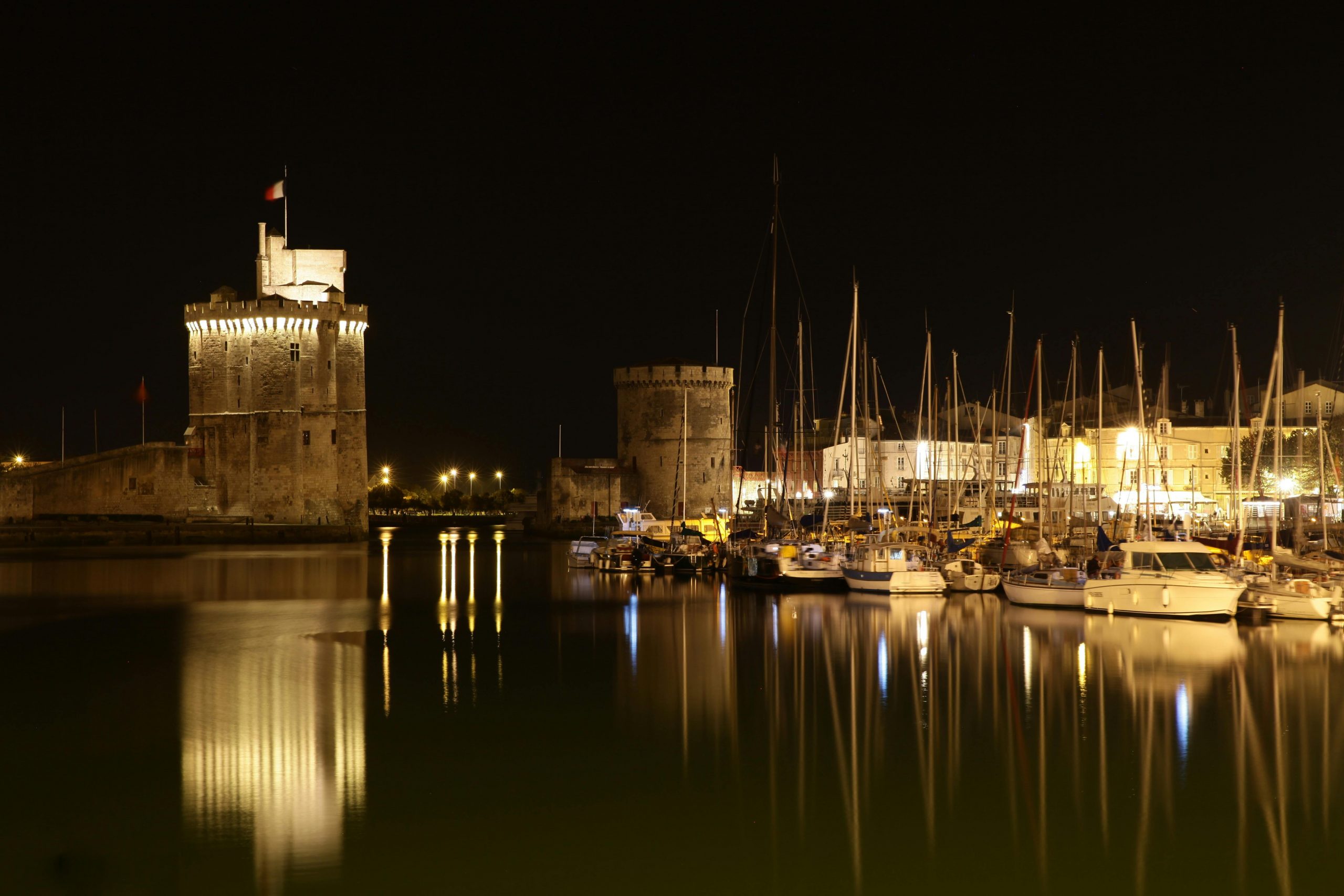 hotographie nocturne du port de La Rochelle, avec ses tours historiques illuminées se reflétant sur l'eau.